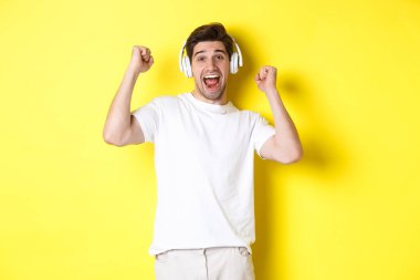 Heyecanlı yakışıklı adam dans ediyor ve şarkı söylüyor, kulaklıkla müzik dinliyor, sarı arka planda duruyor.