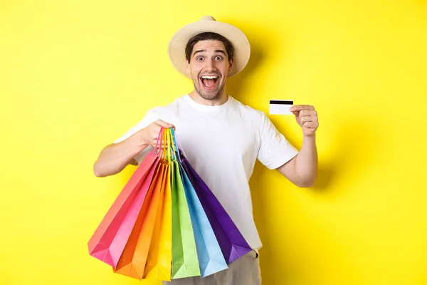 Chico feliz yendo de compras de vacaciones, sosteniendo bolsas de papel y mostrando la tarjeta de crédito, de pie sobre el fondo amarillo —  Fotos de Stock