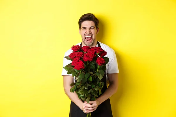 Vicces fiatal eladó fekete kötényben kezében csokor rózsa, virágárus nevet és áll a sárga háttér — Stock Fotó