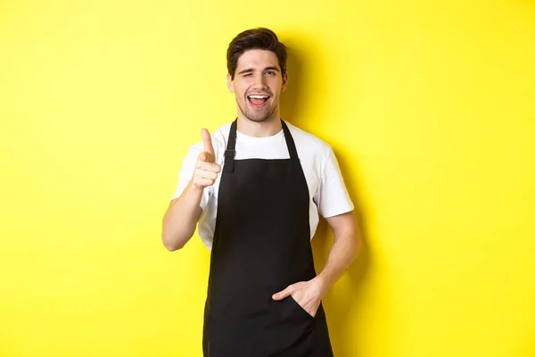 Barista guapo en delantal negro bienvenida a los huéspedes a la cafetería, apuntando con el dedo y guiño, de pie sobre el fondo amarillo — Foto de Stock