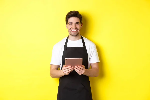 Офіціант в чорному фартусі приймає замовлення, тримає цифровий планшет і посміхається дружній, стоїть над жовтим тлом — стокове фото