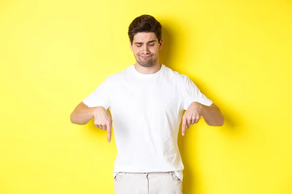 Sceptyczny młody człowiek w białej koszulce, wskazujący i patrzący w dół zdenerwowany, niepochwalający i nielubiący produktu, stojący na żółtym tle — Zdjęcie stockowe