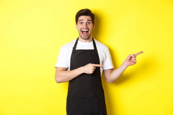 Σερβιτόρος με μαύρη ποδιά που δείχνει τα δάχτυλα δεξιά, δείχνει διαφήμιση και χαμογελά ενθουσιασμένος, στέκεται πάνω σε κίτρινο φόντο — Φωτογραφία Αρχείου