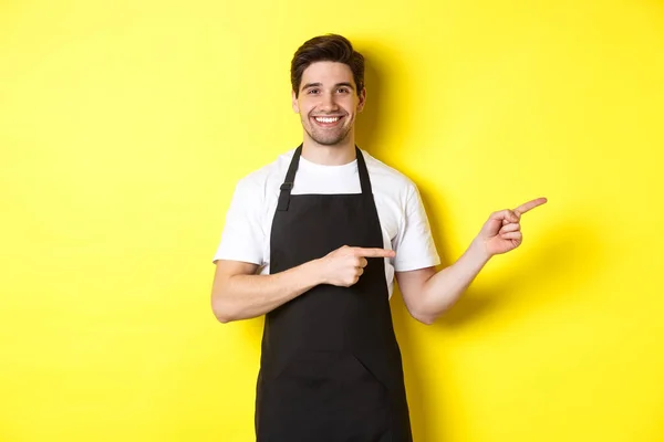Дружній офіціант, який вказує пальці праворуч, показує ваш логотип або рекламну пропозицію, одягнений у чорний фартух, що стоїть на жовтому тлі — стокове фото