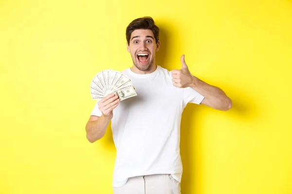 Emocionado hombre sosteniendo el dinero, mostrando el pulgar hacia arriba en la aprobación, consiguió crédito o préstamo, de pie sobre fondo amarillo — Foto de Stock