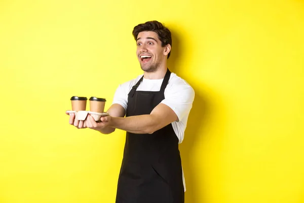 Amistoso barista en delantal negro dando orden de comida para llevar, sosteniendo dos tazas de café y sonriendo, de pie sobre fondo amarillo — Foto de Stock