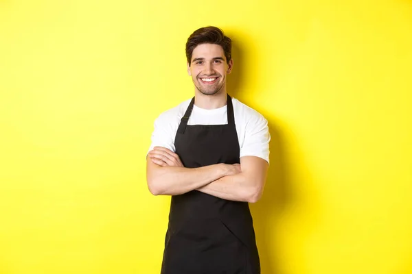 Garçom sorridente masculino em avental preto de pé confiante, braços cruzados no peito contra fundo amarelo — Fotografia de Stock