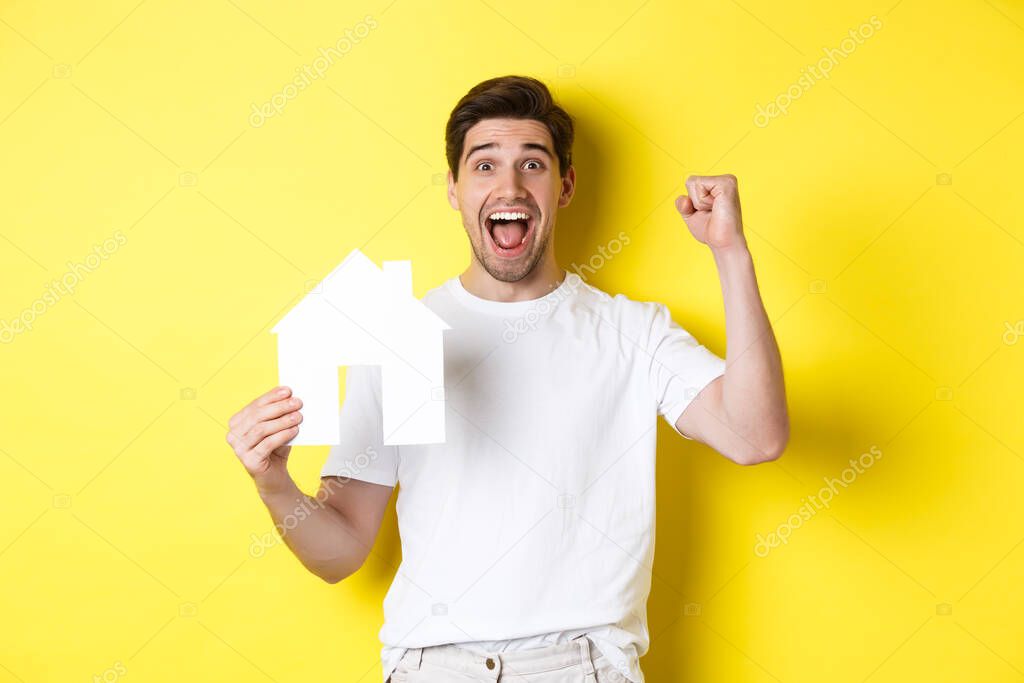 Real estate concept. Cheerful man showing paper house model and making fist pump, paid mortgage, yellow background