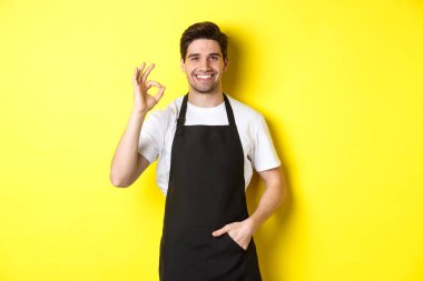 Siyah önlüklü, gülümseyen genç barista, tamam işareti gösteriyor, kahve ya da restoran öneriyor, sarı arka planda duruyor.