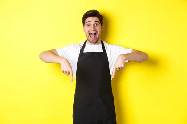 Pelayan, barista warung kopi dengan celemek hitam menunjuk ke bawah, tampak kagum, berdiri di atas latar belakang kuning — Stok Foto