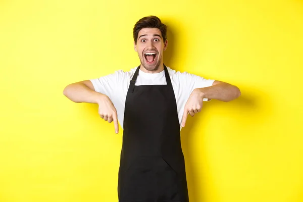 Ober, koffieshop barista in zwarte schort wijzende vingers naar beneden, kijken verbaasd, staande over gele achtergrond — Stockfoto