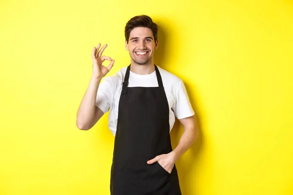 Jonge glimlachende barista in zwart schort toont goed teken, aanbevelen koffieshop of restaurant, staande over gele achtergrond — Stockfoto