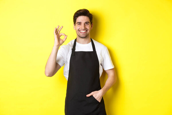 Young smiling barista in black apron showing okay sign, recommending coffee shop or restaurant, standing over yellow background