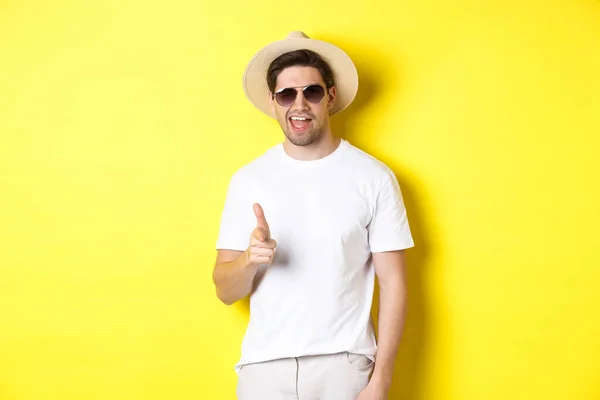 Selvsikker og frekk fyr på ferie som flørter med deg, peker finger mot kamera og blunker, bruker sommerhatt med solbriller, gul bakgrunn – stockfoto