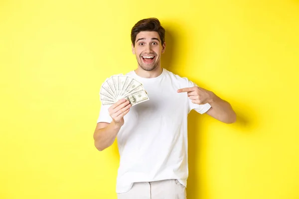 Feliz chico guapo apuntando con el dedo al dinero, concepto de crédito y préstamo, de pie sobre el fondo amarillo — Foto de Stock