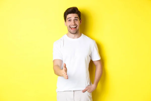 Vriendelijke man begroet u met handdruk, glimlachend geamuseerd, zeggen hallo, staande over gele achtergrond — Stockfoto