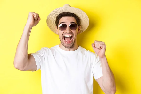 Concepto de turismo y vacaciones. Feliz turista masculino celebrando sus vacaciones, levantando las manos y gritando de alegría, usando gafas de sol con sombrero de paja — Foto de Stock