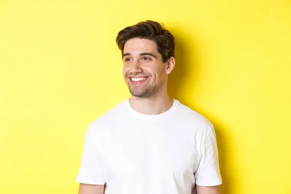 Nære på en kjekk skjeggete mann i hvit t-skjorte som smiler, ser til venstre på kopirommet, står mot gul bakgrunn – stockfoto