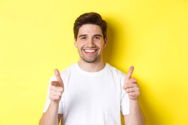 Kendine güvenen adam kamerayı işaret ediyor ve gülümsüyor, seni övüyor, sarı arka planda duruyor.