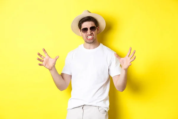 Туризм, путешествия и праздники концепции. Дерзкий молодой человек в отпуске, показывает что-то большое и сжимающиеся зубы, стоя в солнцезащитных очках и летней шляпе — стоковое фото