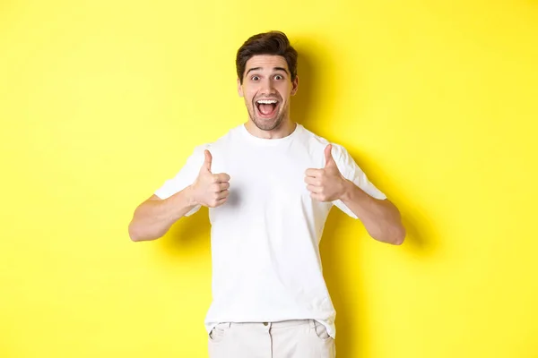 Opgewonden knappe man met duimen omhoog, goedkeuren en ja zeggen, staande over gele achtergrond — Stockfoto