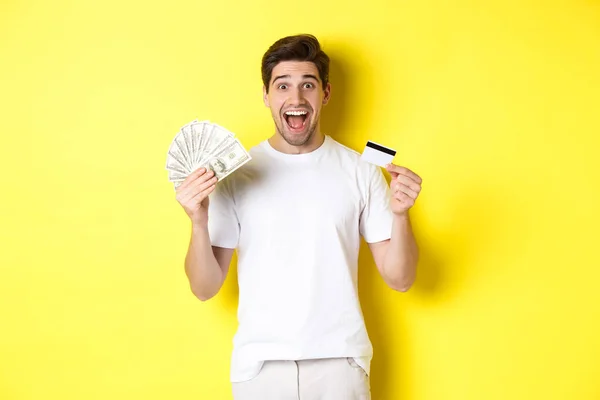 Hombre emocionado listo para ir de compras el viernes negro, sosteniendo dinero y tarjeta de crédito, de pie sobre fondo amarillo — Foto de Stock