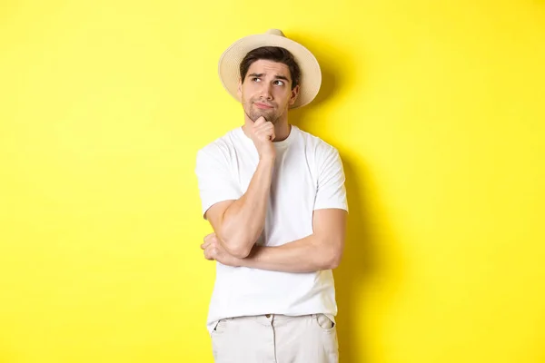 Концепция туризма и лета. Задумчивый турист размышляет, смотрит в левый верхний угол и думает, стоя в соломенной шляпе и белой футболке на желтом фоне — стоковое фото