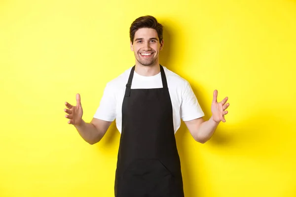 Garçom sorridente em avental preto segurando seu logotipo ou caixa, espalhe as mãos como se carregasse algo grande, de pé sobre o fundo amarelo — Fotografia de Stock