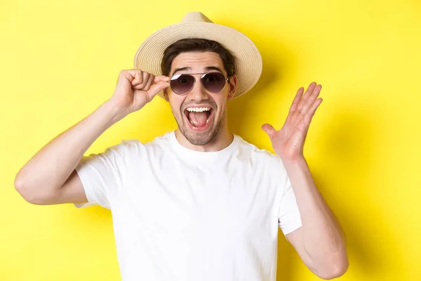 Concepto de turismo y vacaciones. Primer plano del hombre sorprendido gritando de alegría, disfrutando de las vacaciones, usando gafas de sol con sombrero de verano, fondo amarillo — Foto de Stock