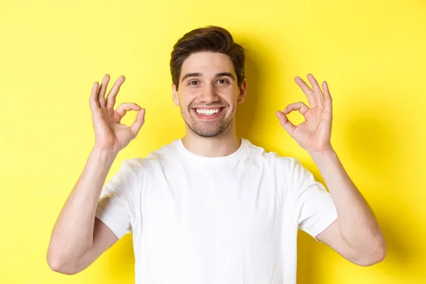 Nære på en kjekk ung mann som viser ok tegn, godkjenner og er enig, smilende fornøyd, står over gul bakgrunn – stockfoto