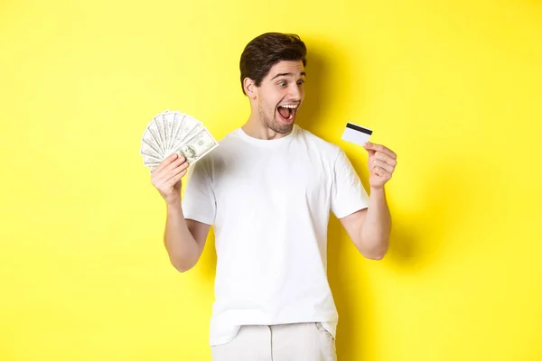 Chico alegre mirando la tarjeta de crédito, sosteniendo el dinero, concepto de crédito bancario y préstamos, de pie sobre fondo amarillo — Foto de Stock