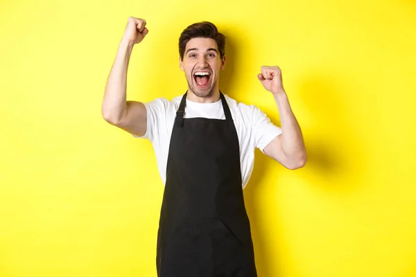 Happy barista merayakan kemenangan, mengangkat tangan dan berteriak-teriak untuk sukacita, mengenakan celemek hitam, seragam toko, berdiri melawan latar belakang kuning — Stok Foto