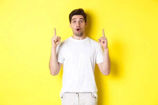 Förvånad snygg kille i vit t-shirt, pekar finger upp, intresserad av reklam, står mot gul bakgrund — Stockfoto