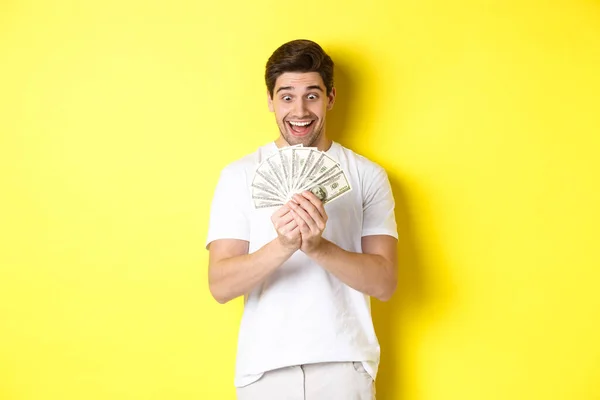 Hombre feliz mirando el dinero y sonriendo emocionado, ganando el premio, consiguió préstamo bancario, de pie sobre fondo amarillo — Foto de Stock