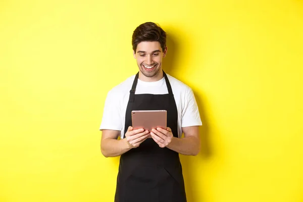 Продавець в чорному фартусі, дивлячись на екран цифрових планшетів, посміхаючись задоволений, стоїть над жовтим тлом — стокове фото