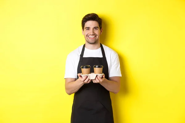 Красивий чоловічий бариста, який подає каву на винос і посміхається, приносячи порядок, стоїть в чорному фартуху на жовтому тлі — стокове фото