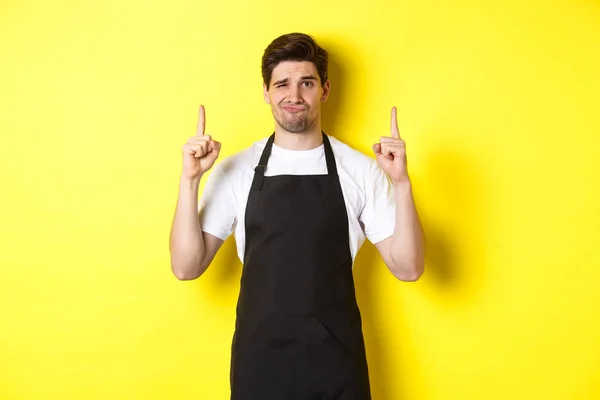Неприємний і скептичний офіціант в чорному фартуху, який вказує пальці вгору, виражає неприязнь і байдужість, стоїть на жовтому тлі — стокове фото
