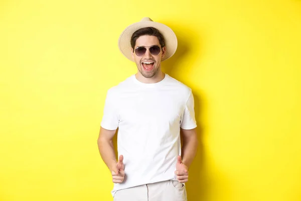 自信と生意気な男オン休暇あなたといちゃつく,カメラとウインクで指を指して,サングラスで夏の帽子を身に着けています,黄色の背景 — ストック写真