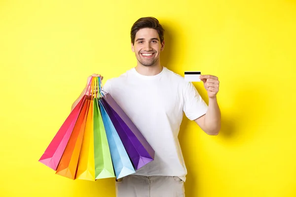 Young man shopping for holidays, holding paper bags and recommending bank credit card, standing over yellow background — Stock Photo, Image