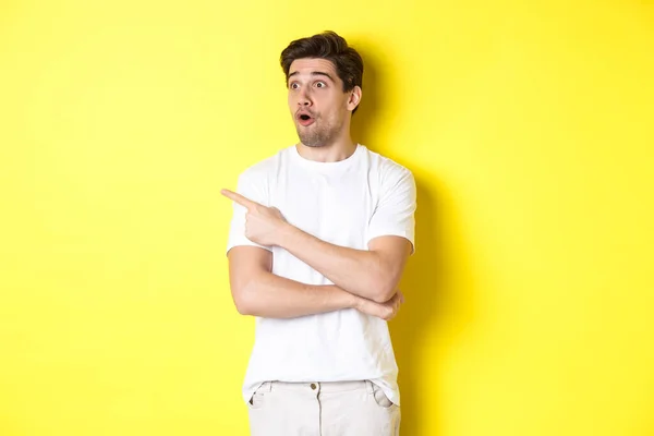 Homem impressionado em t-shirt branca, olhando e apontando o dedo deixado para promo, confira anúncio, de pé contra fundo amarelo — Fotografia de Stock