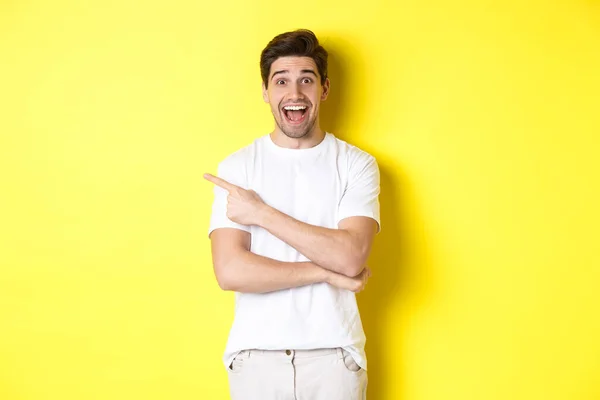 Szczęśliwy człowiek wskazujący palcem w lewo, pokazujący reklamę na przestrzeni kopiowania, uśmiechnięty rozbawiony, stojący w białych ubraniach na żółtym tle — Zdjęcie stockowe