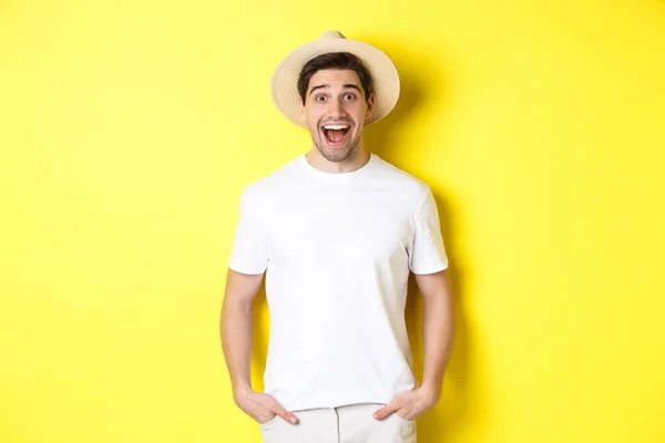 Overrasket mann turist i stråhatt ser glad ut, reagerer forbløffet på reiselivsreklame, står over gul bakgrunn – stockfoto
