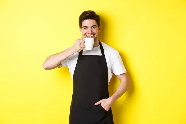 Kafe barista minum secangkir kopi dan tersenyum, mengenakan celemek hitam, berdiri di atas latar belakang kuning — Stok Foto