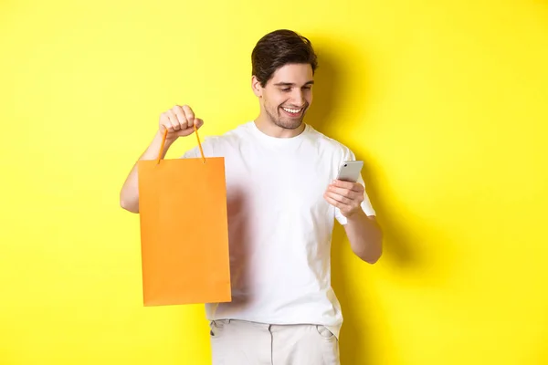 Concepto de descuentos, banca en línea y reembolso. Chico feliz mostrando bolsa de compras y buscando satisfecho en la pantalla móvil, fondo amarillo — Foto de Stock