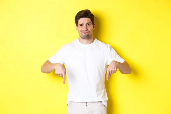 Jovem infeliz sorrindo, apontando os dedos para baixo na propaganda, decepcionado com o produto, de pé sobre fundo amarelo — Fotografia de Stock