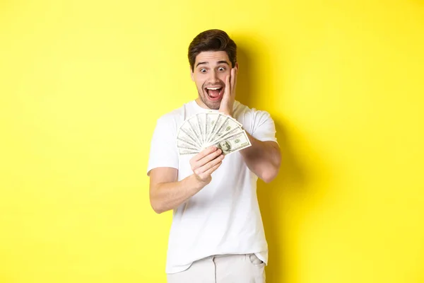 Hombre mirando sorprendido por el dinero, ganar premio en efectivo, de pie sobre fondo amarillo — Foto de Stock