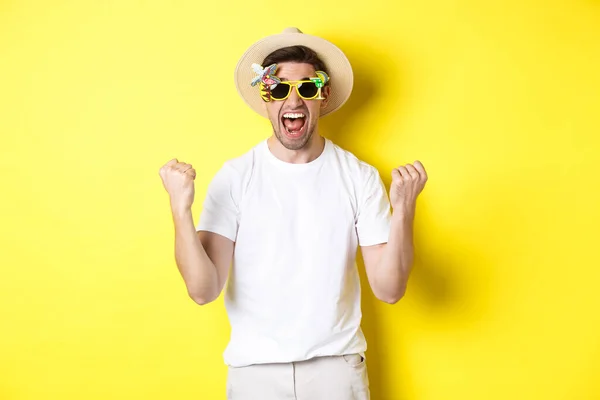 Chico feliz yendo de vacaciones, ganando o celebrando, usando sombrero de verano y gafas de sol. Turista mirando emocionado, de pie sobre fondo amarillo — Foto de Stock