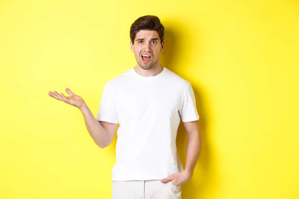 Verward en geschokt man klagen, het opheffen van een hand en kijken lastig, staan in de buurt van gele kopieerruimte — Stockfoto