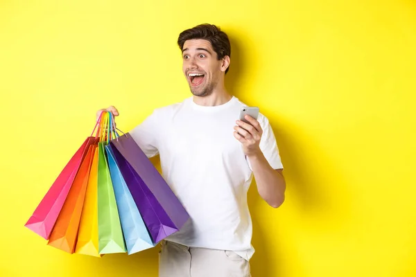 Concepto de banca móvil y reembolso. Hombre feliz mirando sorprendido, sosteniendo bolsas de compras y teléfono inteligente, fondo amarillo —  Fotos de Stock