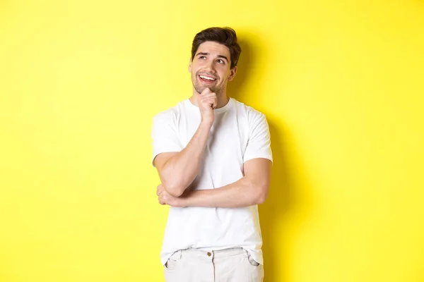 Portrait of smiling man looking thoughtful at upper left corner, choosing something, have an idea, standing against yellow background — Stock Photo, Image